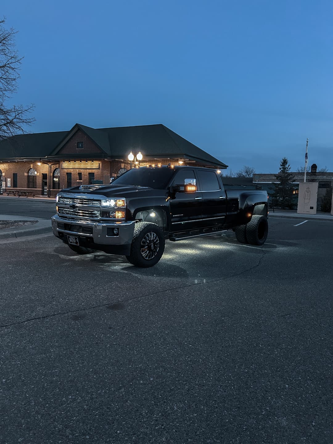 Black lifted pickup truck parked in front of a building during dusk with glowing headlights.