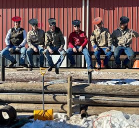 Group of five people in hats sitting on a fence at a farm, snow and tools in foreground.