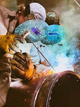 Welders using a welding torch on metal, sparks flying during fabrication process.