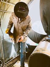 Welder in protective gear working inside a workshop with welding equipment.