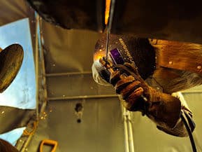 Welder in protective gear working on metal, sparks flying in a workshop environment.