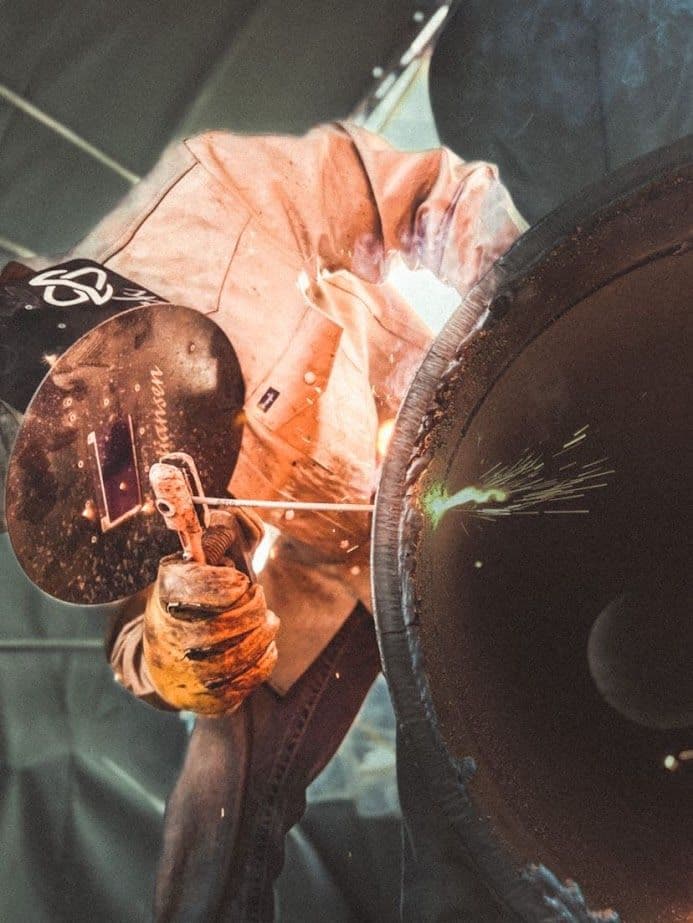 Welder in protective gear performing a weld on a large metal pipe, sparks flying.