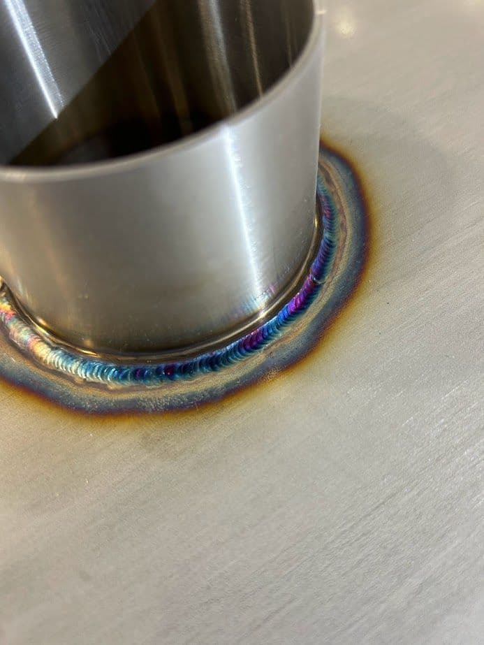Metal weld joint with colorful heat-affected zone around cylindrical stainless steel tube.