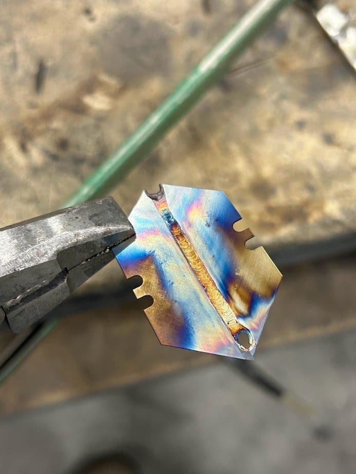 Metal art piece with vibrant colors and welds, held by pliers on a workbench.