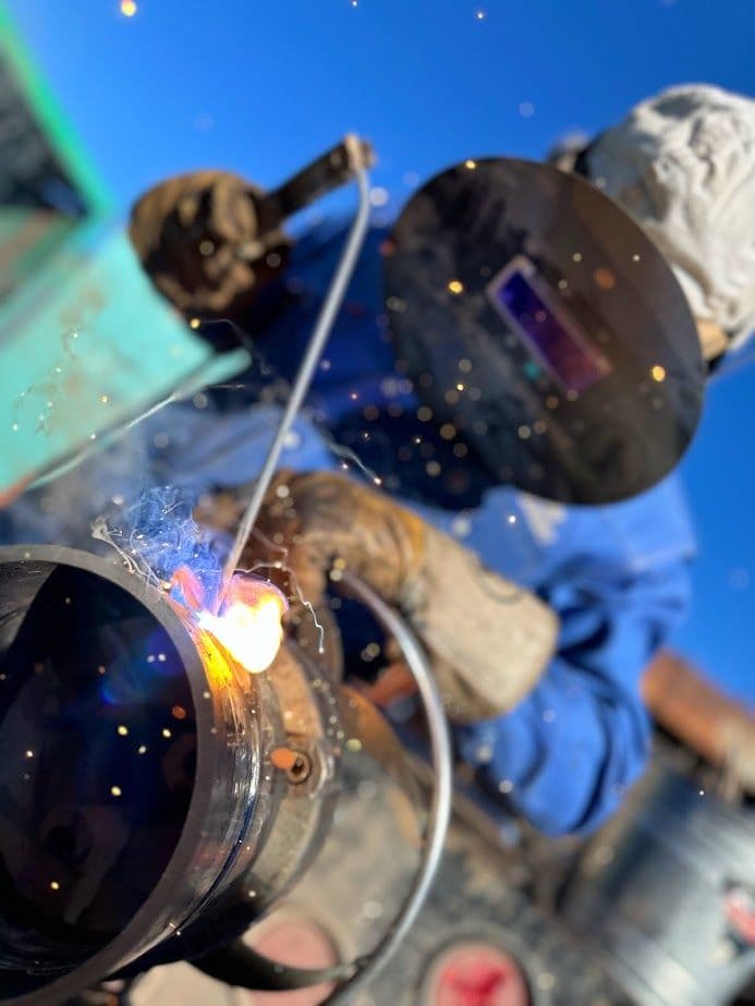 Welder in protective gear using a torch, sparks flying against a blue sky backdrop.
