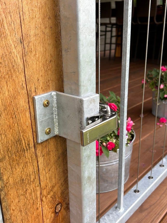 Metal gate latch secured with a padlock, attached to wooden post and surrounded by flowers.