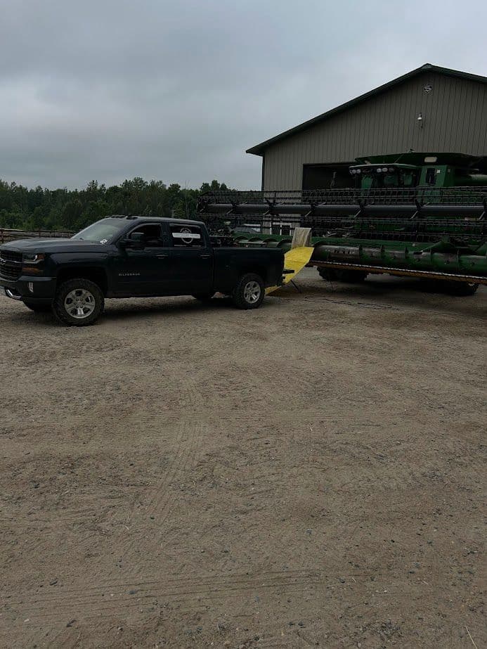 Pickup truck towing a green combine harvester near a storage facility on a gravel road.