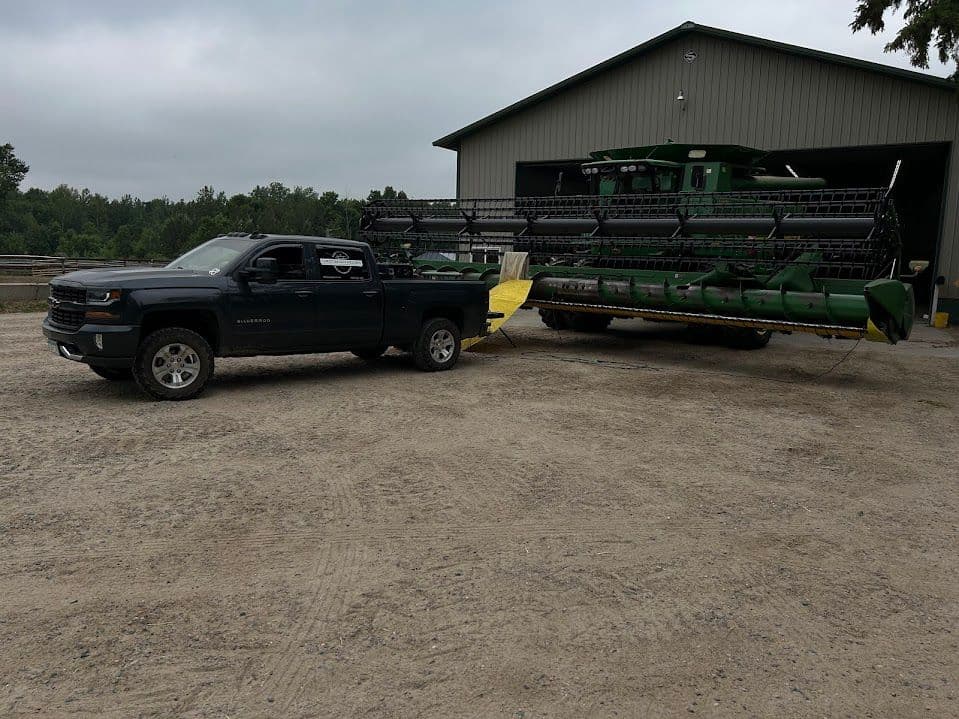 Pickup truck towing a large agricultural harvester near a barn on a gravel lot.