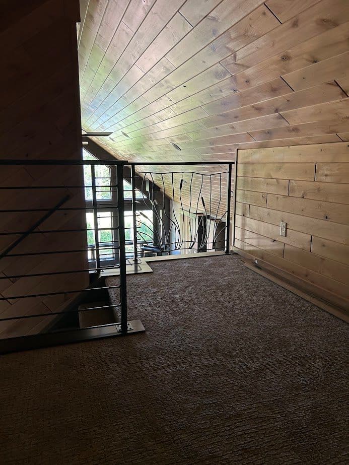 Cozy loft space with wooden walls, metal railing, and carpeted floor overlooking a scenic view.