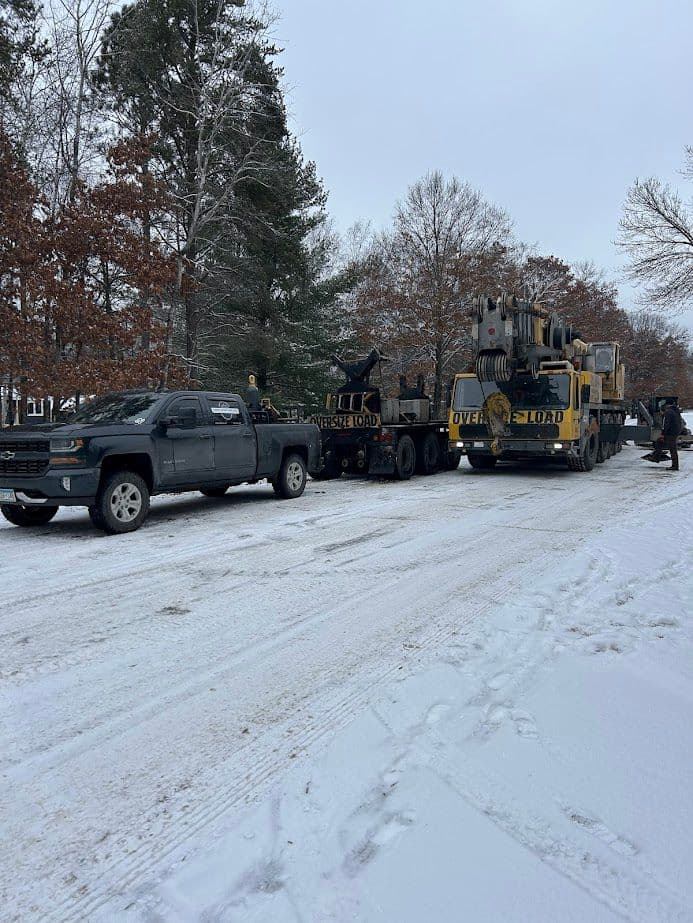 Truck and heavy machinery on a snowy road, surrounded by trees in winter landscape.