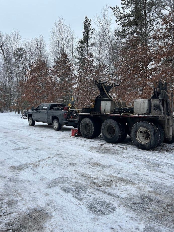 Truck and trailer parked in snowy forest with bare trees in winter landscape.