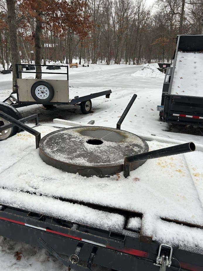 Metal round platform with handles on a snowy trailer, surrounded by trees. Winter scene.