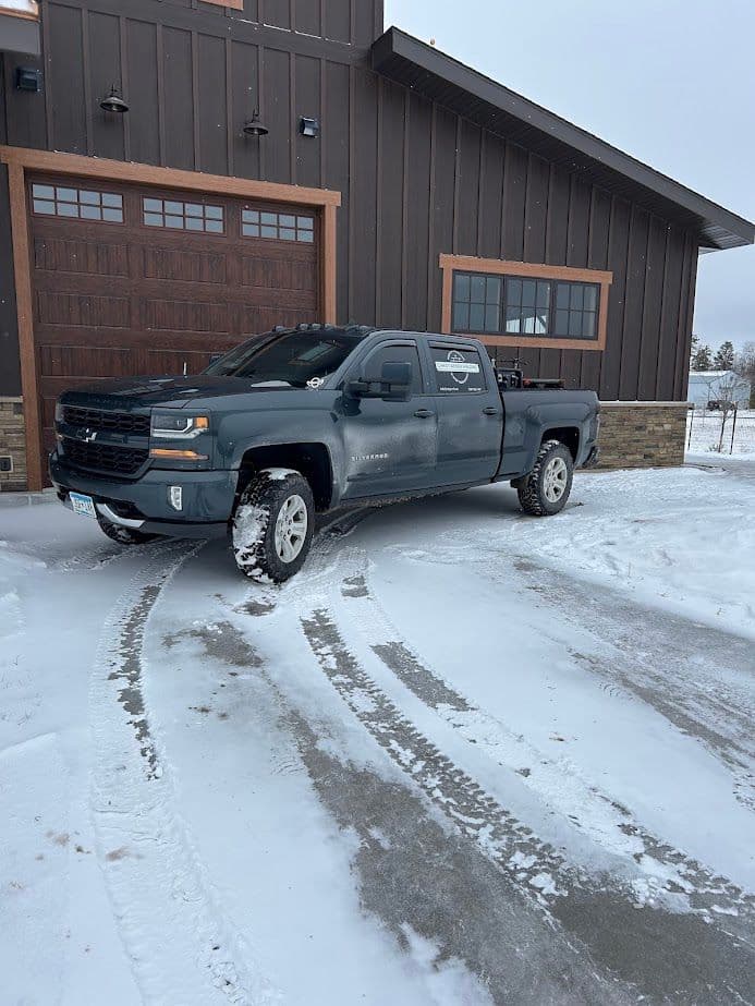 Chevrolet pickup truck parked in snowy driveway next to modern wooden garage.