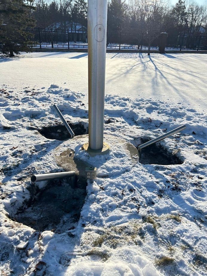 Snow-covered ground with a flagpole and four metal supports in concrete holes.