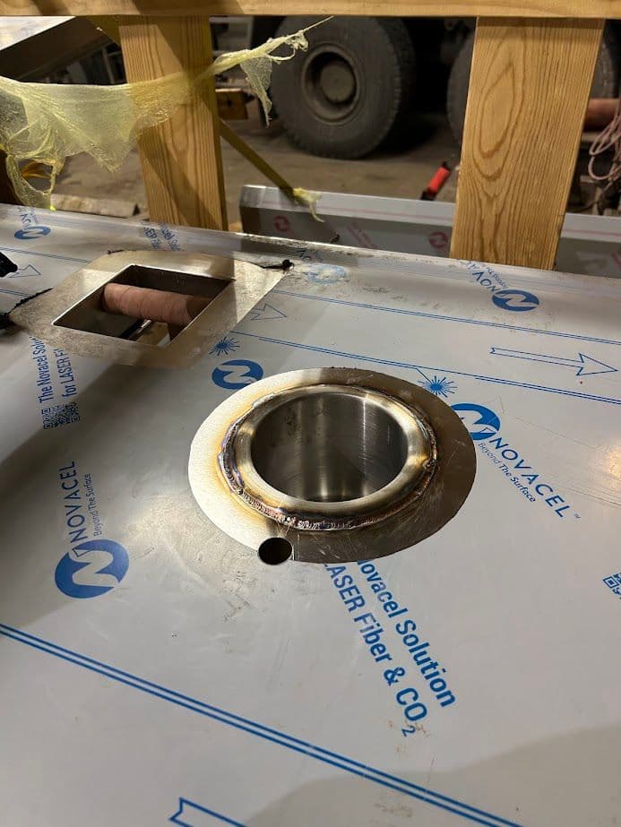 Welded metal component with circular cutout on a workshop table, with protective film still on.