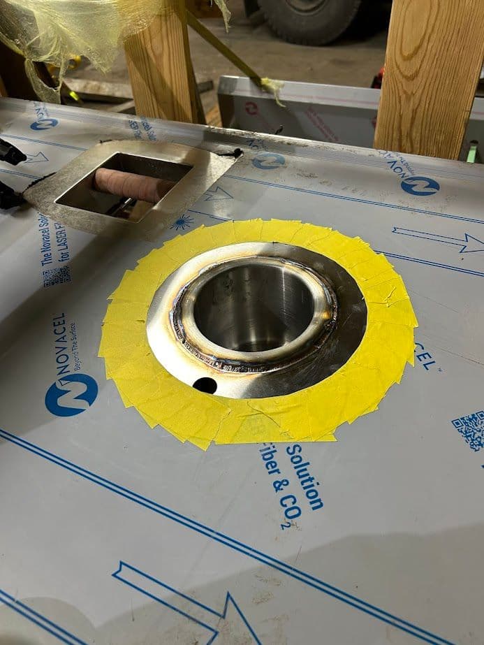 Stainless steel circular cutout with yellow tape on a metal surface in a workshop setting.