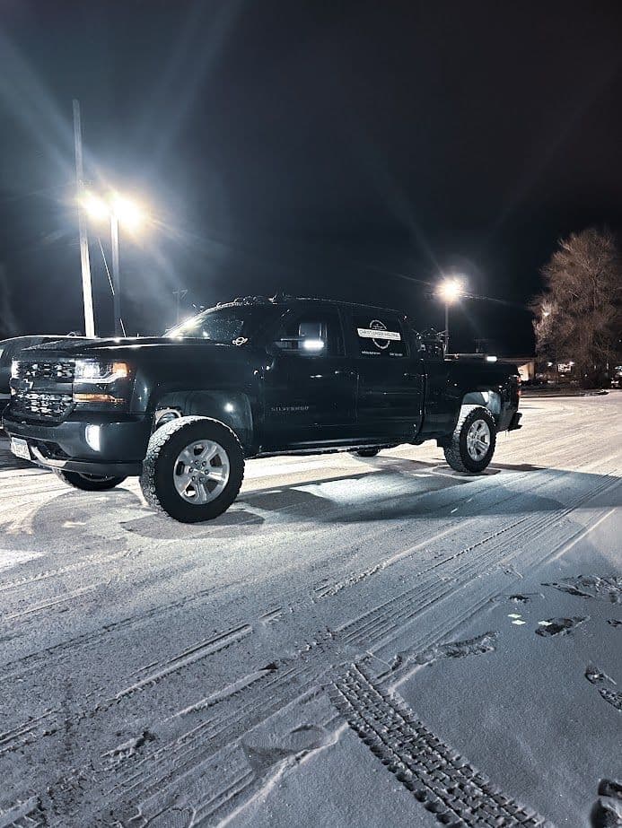 Black pickup truck parked in the snow, lit by bright overhead lights at night.