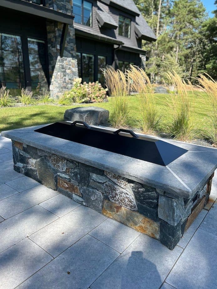 Modern stone firepit with a sleek black cover in a landscaped outdoor setting.