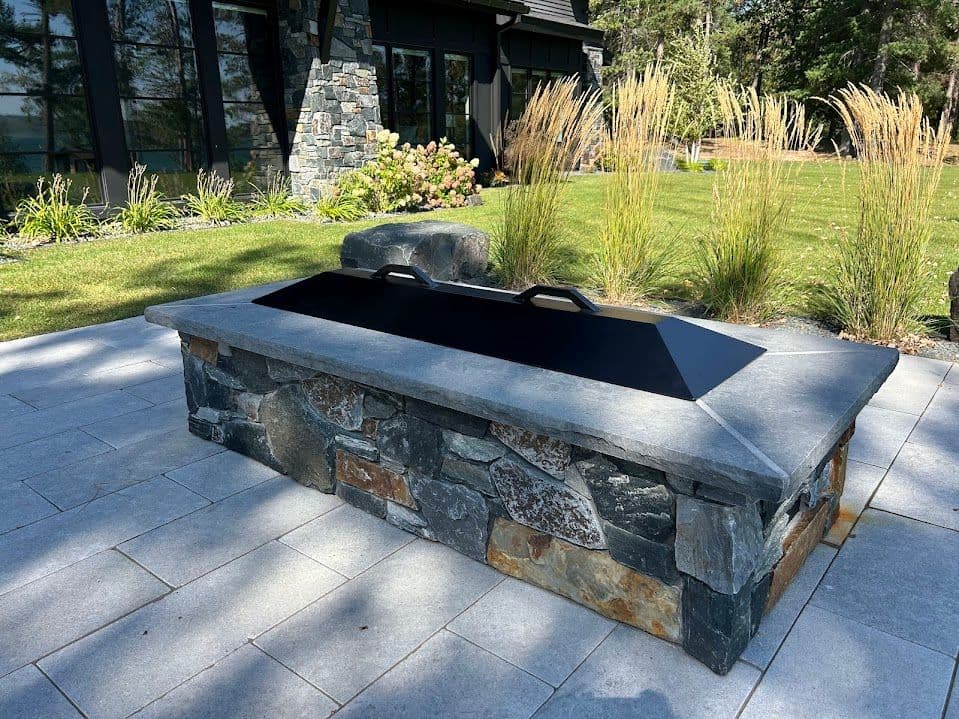 Modern stone fire pit with a black lid in a landscaped outdoor setting.