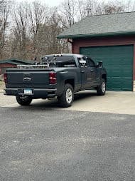 Black Chevrolet truck parked in driveway next to a green garage on a wooded property.