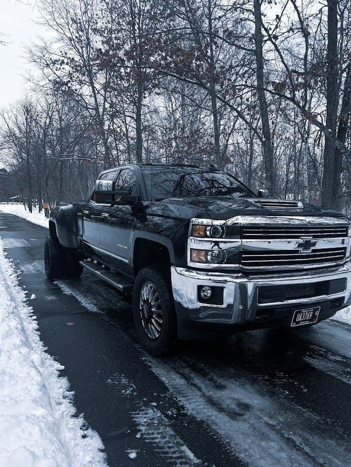 Black Chevrolet truck parked on snowy road surrounded by trees in winter scene.