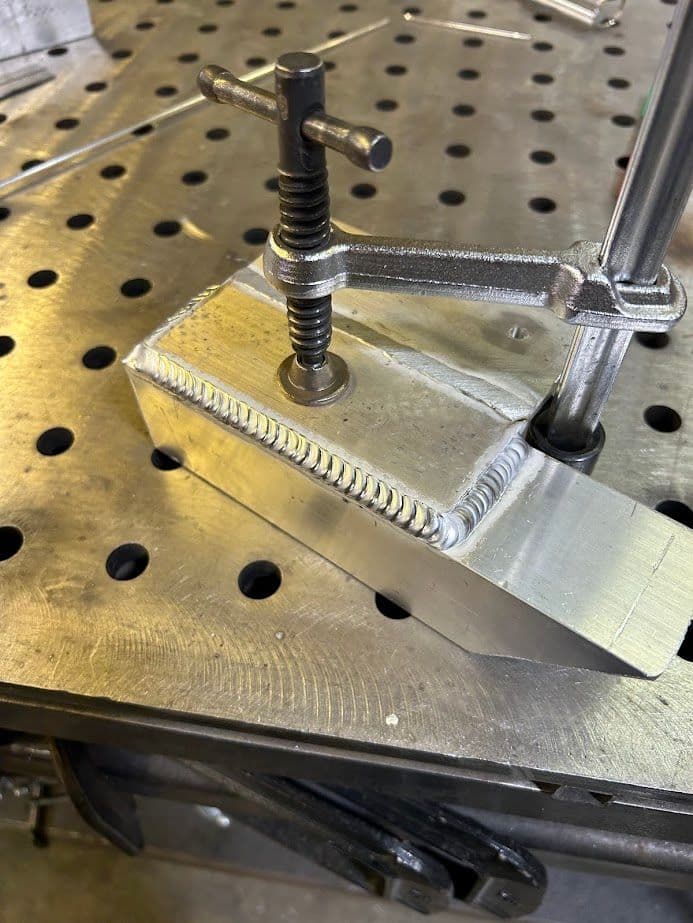 Aluminum workpiece with threaded clamp and welded seam on a metal workbench.