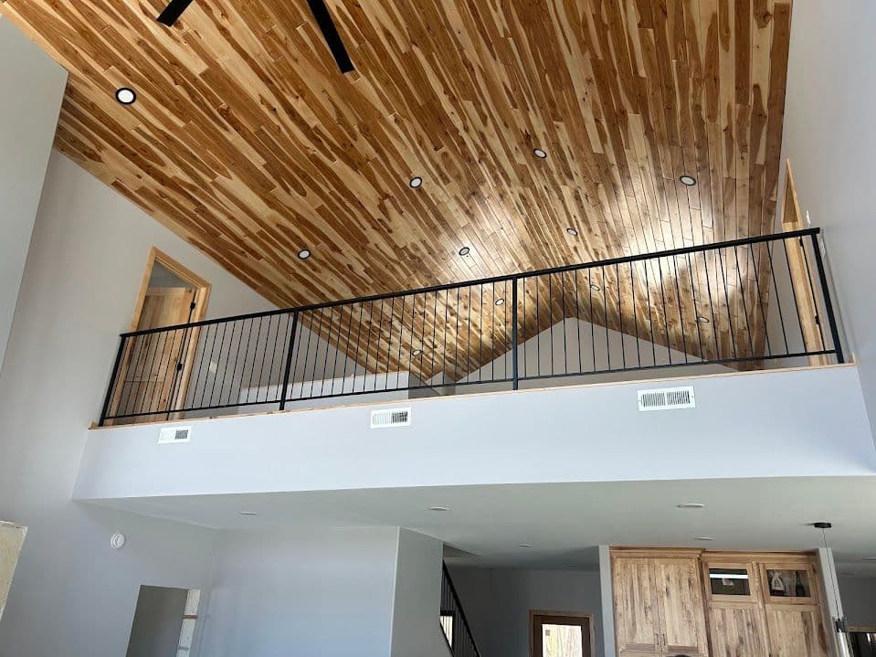 Bright interior with wooden vaulted ceiling and black railing overlooking a spacious living area.