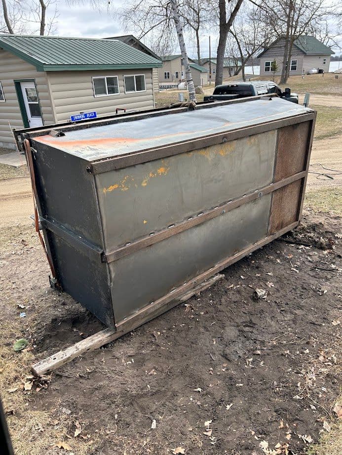 Abandoned metal dumpster on a dirt area near residential buildings in a rural setting.