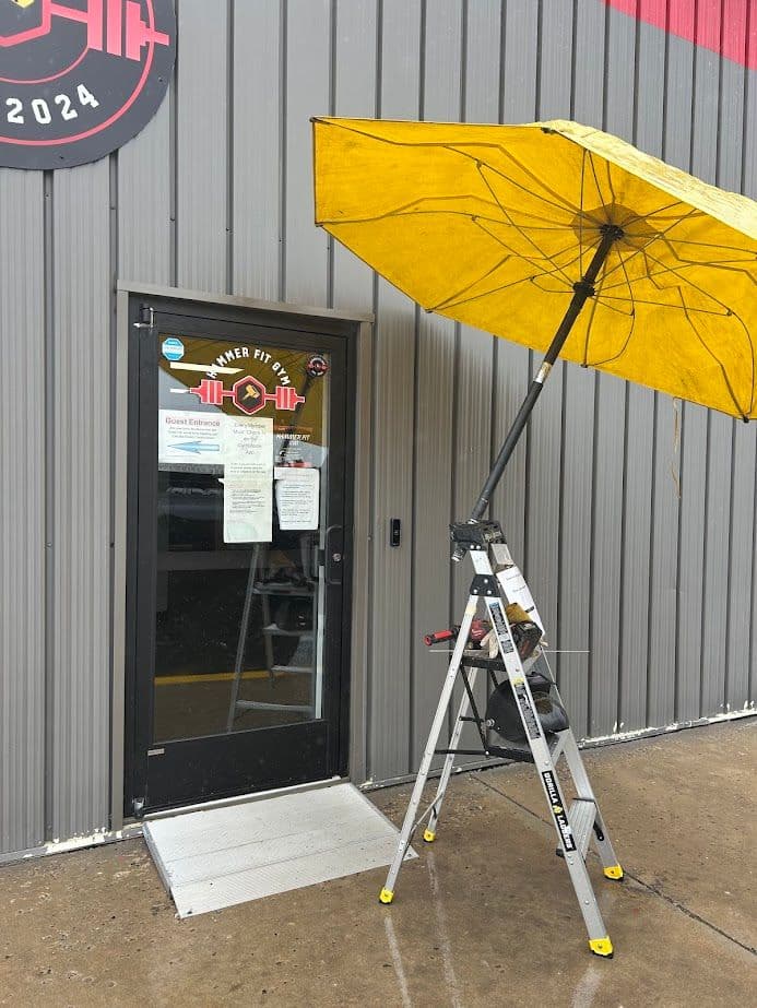 Entrance of Hammer Fit gym with a yellow umbrella covering the door, ladder positioned nearby.
