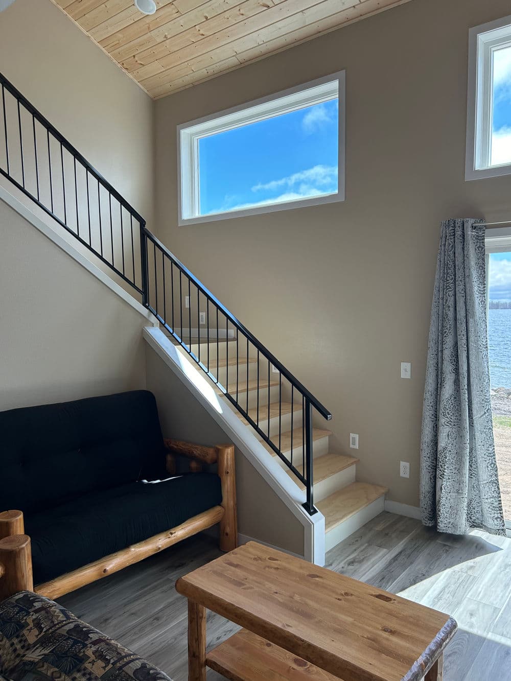 Modern living room with stairs, large windows, and scenic view by the water. Cozy decor elements.