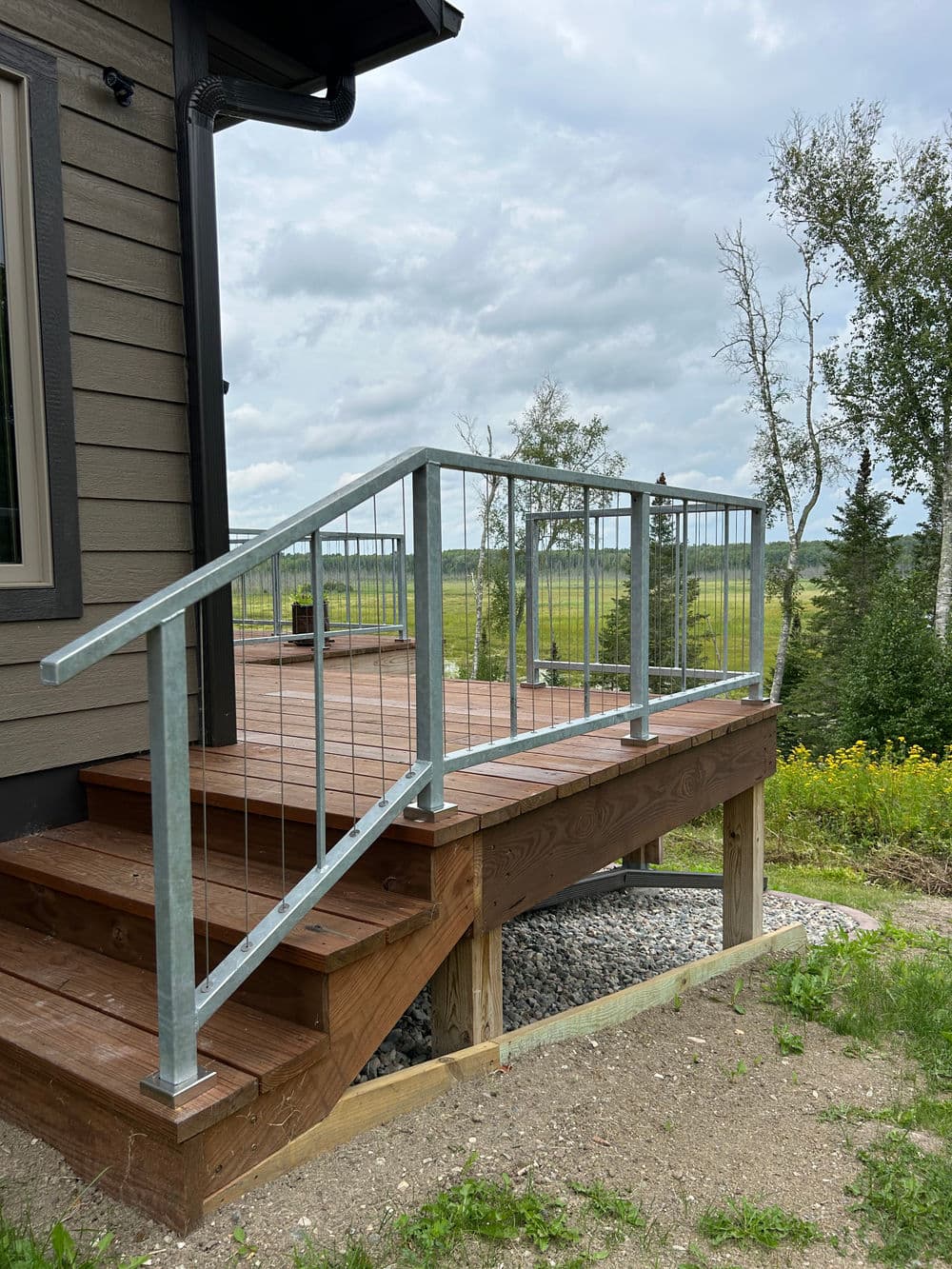 Metal railing on wooden deck with steps, surrounded by greenery and cloudy sky.