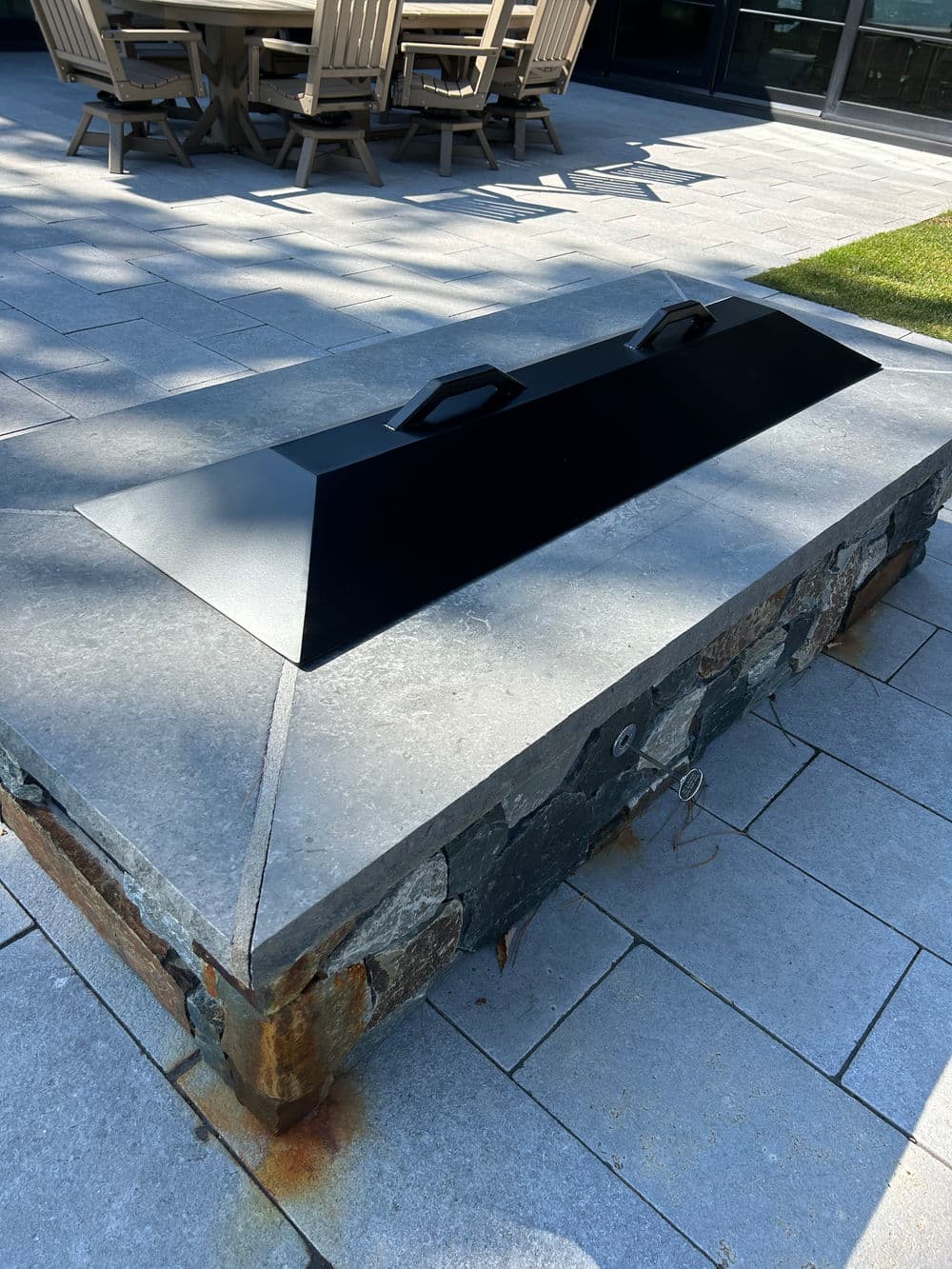 Modern outdoor fire pit with a sleek black cover on a stone base and surrounding patio furniture.