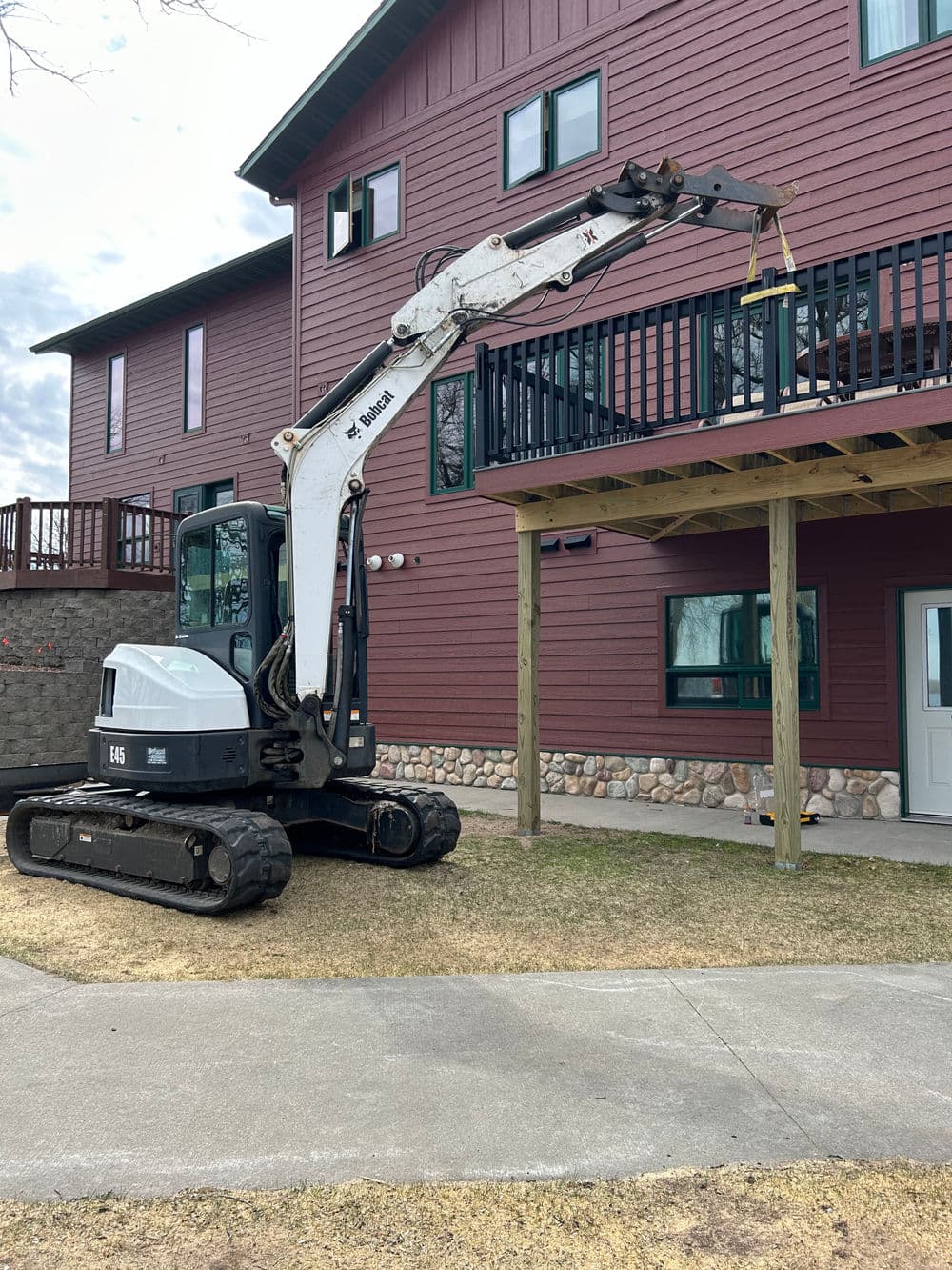 Excavator lifting materials near a residential building with wooden deck.