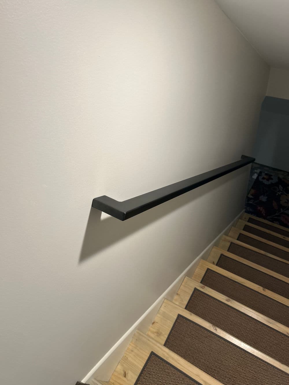Modern stair railing along a beige wall leading down wooden stairs with textured runner.